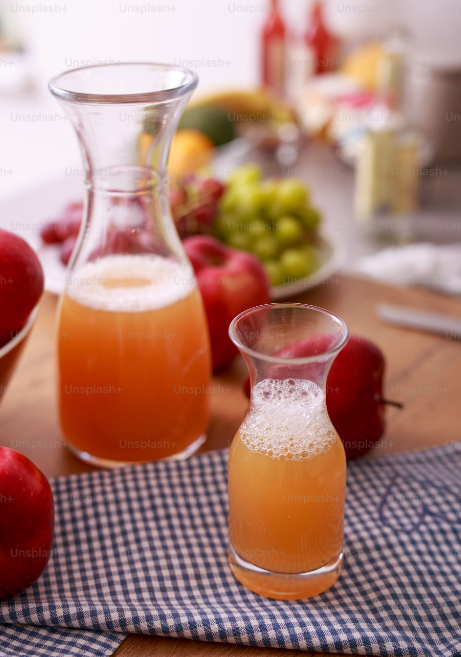 Cette image montre une carafe et un verre de jus de pomme mousseux sur une nappe à carreaux, avec des pommes rouges et des raisins en arrière-plan.