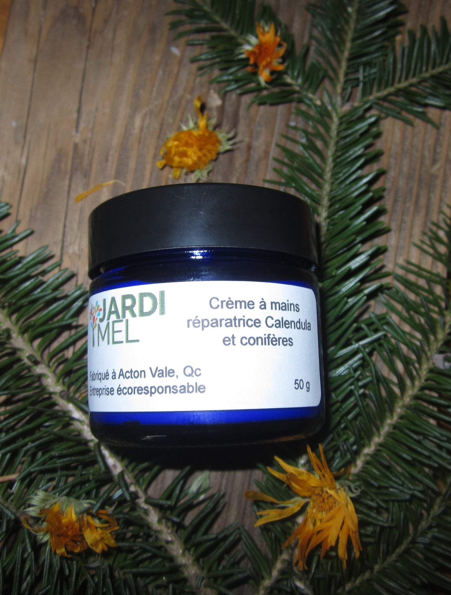 Pot de crème à mains réparatrice de la marque JARDI MEL, entouré de branches de conifères et de fleurs de calendula, fabriqué à Acton Vale, Québec - 50g.