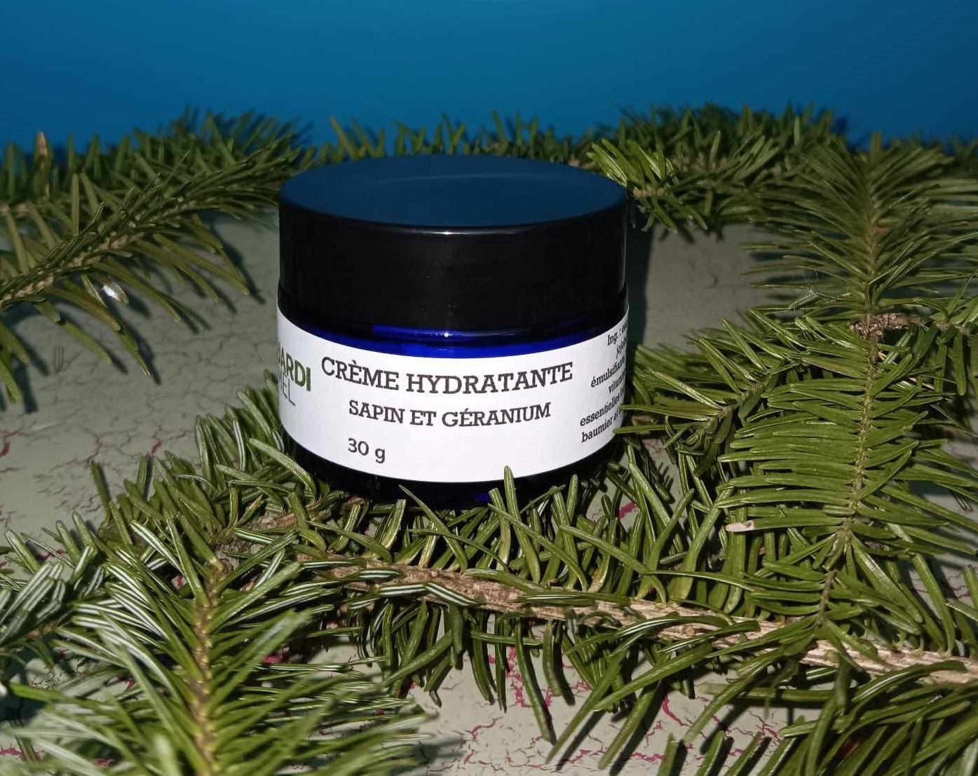 Pot de crème hydratante de 30g "CRÈME HYDRATANTE SAPIN ET GÉRANIUM", posé sur un lit de branches de sapin, avec un couvercle noir et une étiquette bleue mentionnant des ingrédients naturels essentiels et jardimel.com
