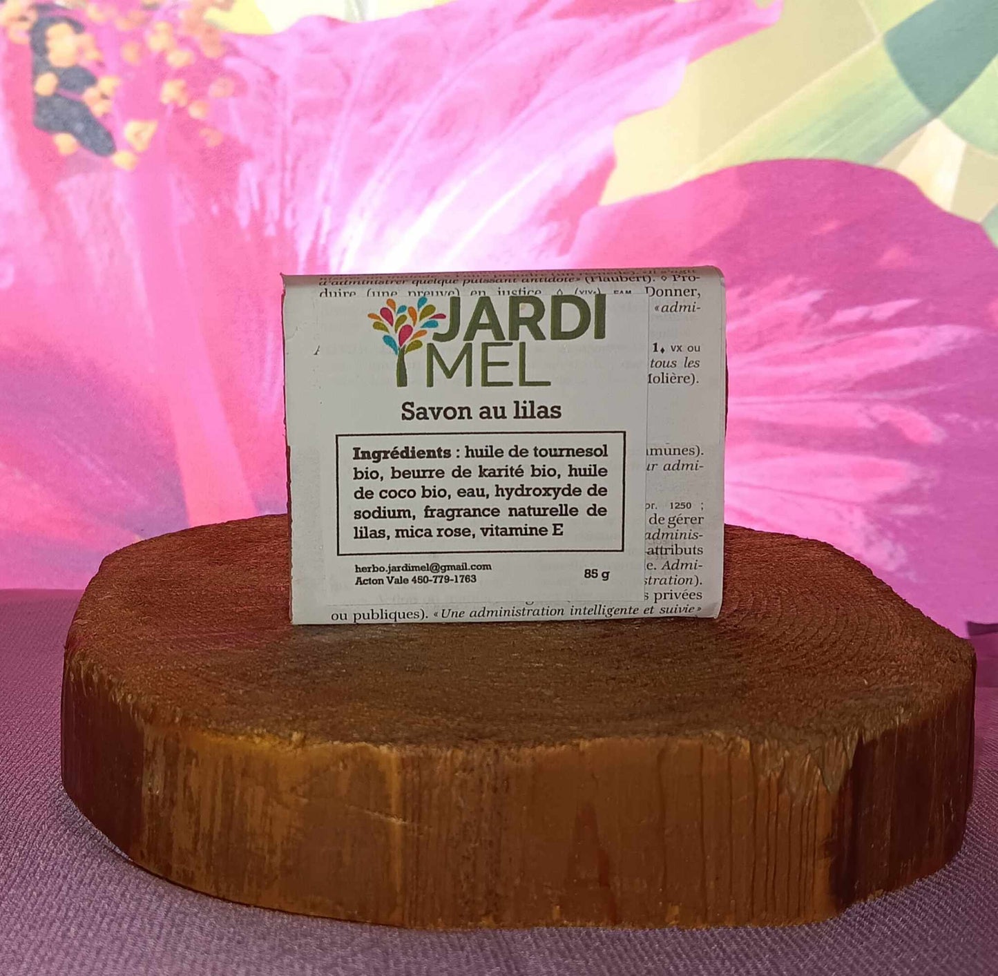 Savon artisanal "Savon au lilas" de la marque Jardi Mel, posé sur une surface en bois avec des fleurs en arrière-plan. Ingrédients : huile de tournesol bio, beurre de karité bio, huile de coco bio, eau, hydroxyde de sodium, fragrance naturelle de lilas, mica rose, vitamine E.