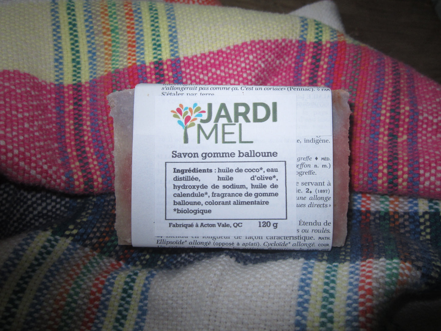 Savon artisanal "Savon gomme balloune" de la marque Jardi Mel, fabriqué à Acton Vale, QC, et pesant 120 g. Les ingrédients biologiques incluent l'huile de coco bio, l'huile d'olive bio, et l'huile de calendule bio. Savon posé sur un tissu coloré à motifs. Étiquette indiquant jardimel.com