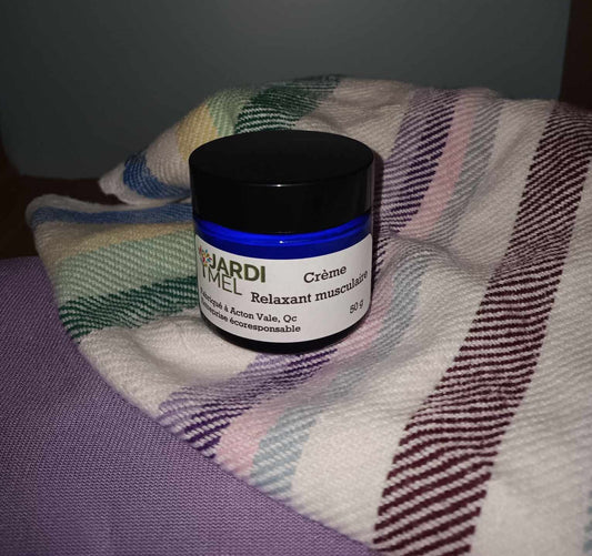 L'image montre un pot de crème relaxante musculaire de la marque Jardi Mel, placé sur une couverture rayée multicolore. Le pot est bleu avec un couvercle noir et une étiquette blanche. L'étiquette indique que le produit est fabriqué à Acton Vale, Qc, et qu'il s'agit d'une entreprise écoresponsable. Le pot contient 50 g de crème.