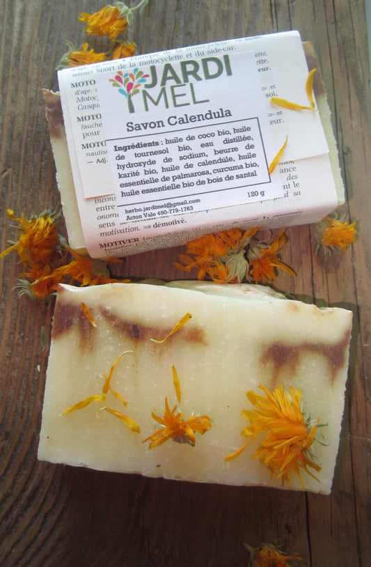 Deux savons artisanaux de Jardi Mel, posés sur une surface en bois et entourés de fleurs de calendula. Le savon emballé indique les ingrédients : huile de coco bio, huile de tournesol bio, beurre de karité bio, huile de calendula, huile essentielle de palmarosa, curcuma bio, huile essentielle de bois de santal. Le savon déballé montre une texture crémeuse avec des pétales de calendula incrustés.