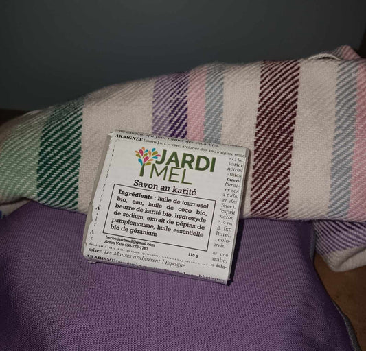 Savon artisanal au karité de la marque Jardi Mel, emballé avec soin et posé sur un tissu coloré à rayures. Les ingrédients naturels incluent l'huile de tournesol bio, l'huile de coco bio, et le beurre de karité aussi biologique. Étiquette avec jardimel.com