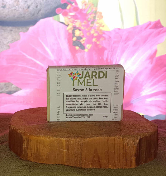 Savon à la rose, de marque Jardi Mel, posée sur une surface en bois, avec une fleur rose en arrière-plan. Ingrédients listés : huile d'olive bio, beurre de karité bio, huile de coco bio, eau distillée, hydroxyde de sodium, huile essentielle de bois de Hô bio, fragrance naturelle de rose, argile rose, vitamine E, et pétales de rose. Étiquette avec jardimel.com