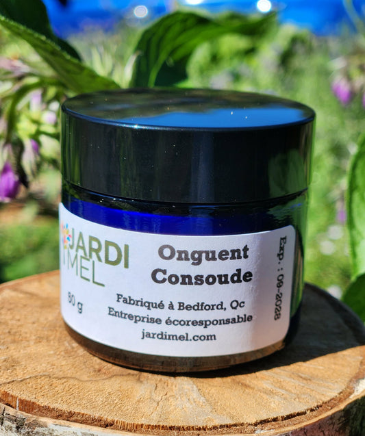 Pot en verre bleu d'onguent de consoude de la marque Jardi Mel, posée sur un rondin de bois naturel devant des fleurs de consoude
