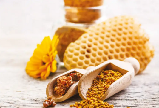 L'image montre des morceaux de propolis dans une cuillère en bois, entourés de produits apicoles comme du pollen, un rayon de miel et un pot de miel.